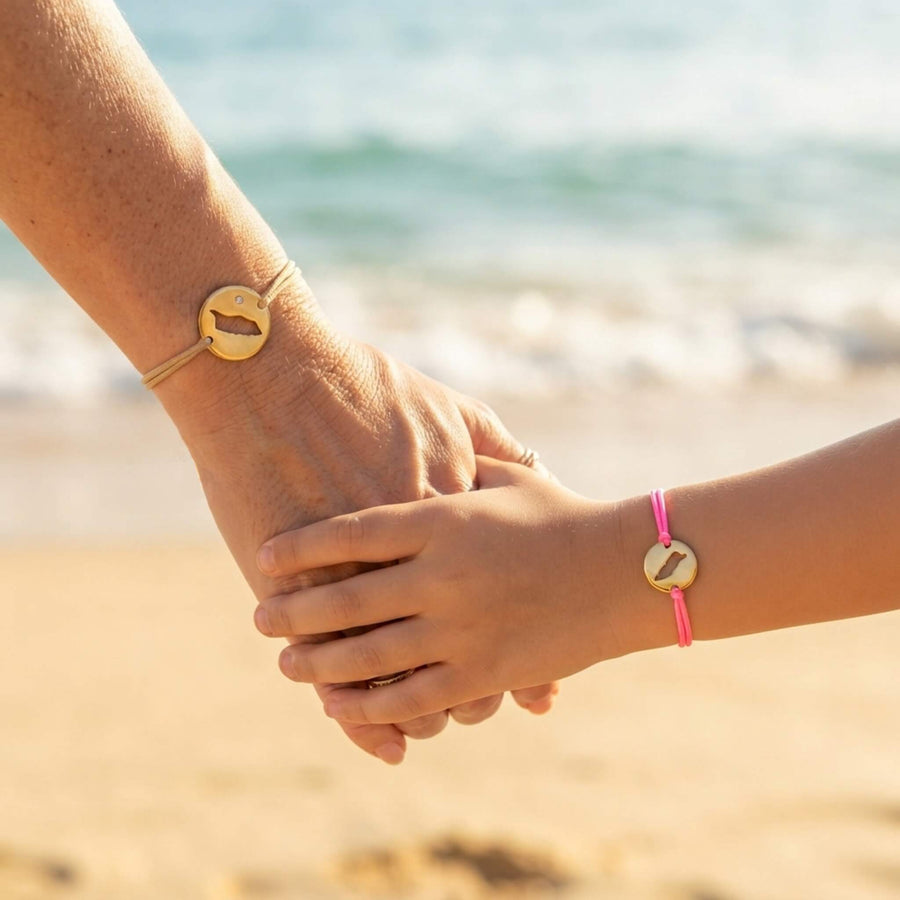 A mother and her child daughter holding hands on a sandy beach, each wearing a thin cord bracelet with a round gold-toned pendant featuring a cut-out map of Aruba.