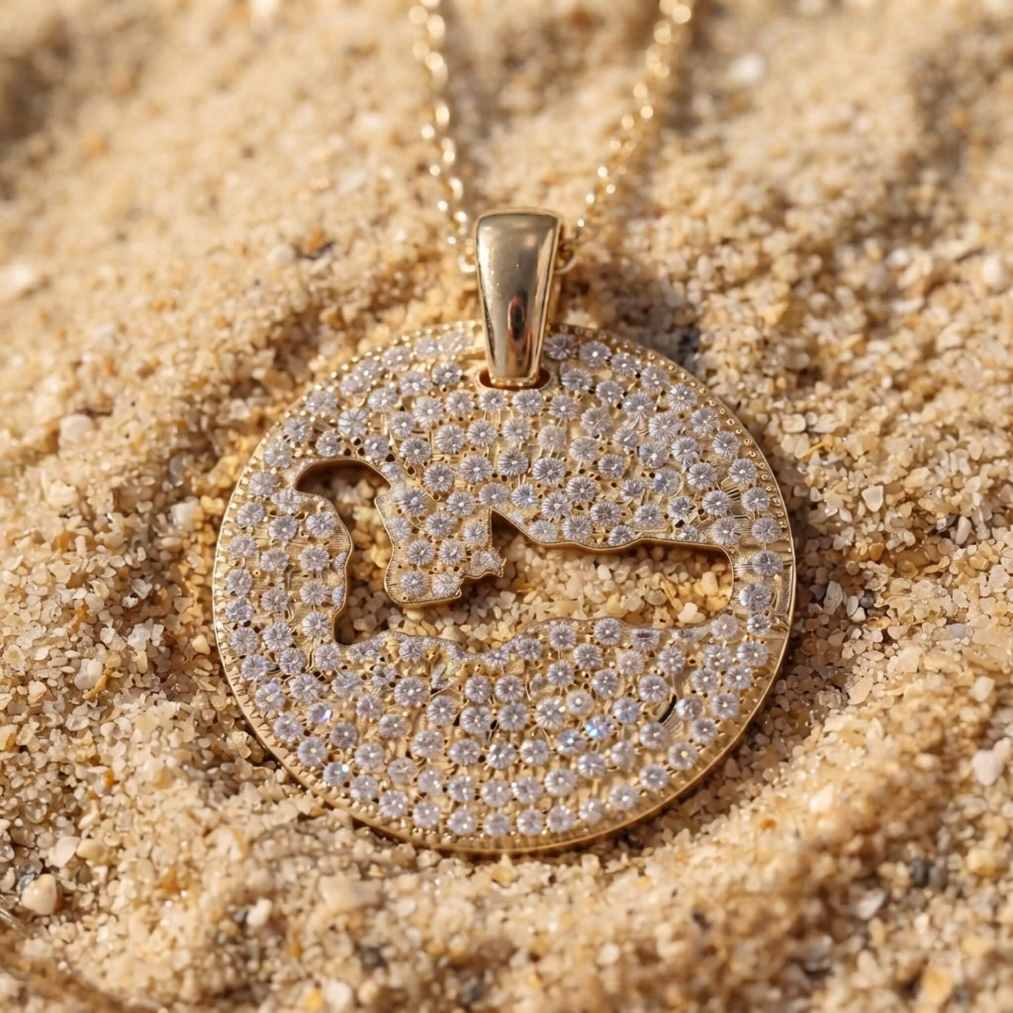 A gold pavé round pendant necklace with a cut-out map of Grand Cayman island, resting on light beige sand.