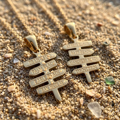 Two DENIZEN gold signpost necklaces with names of places in Aruba engraved, resting on sand with small shells and sea glass.