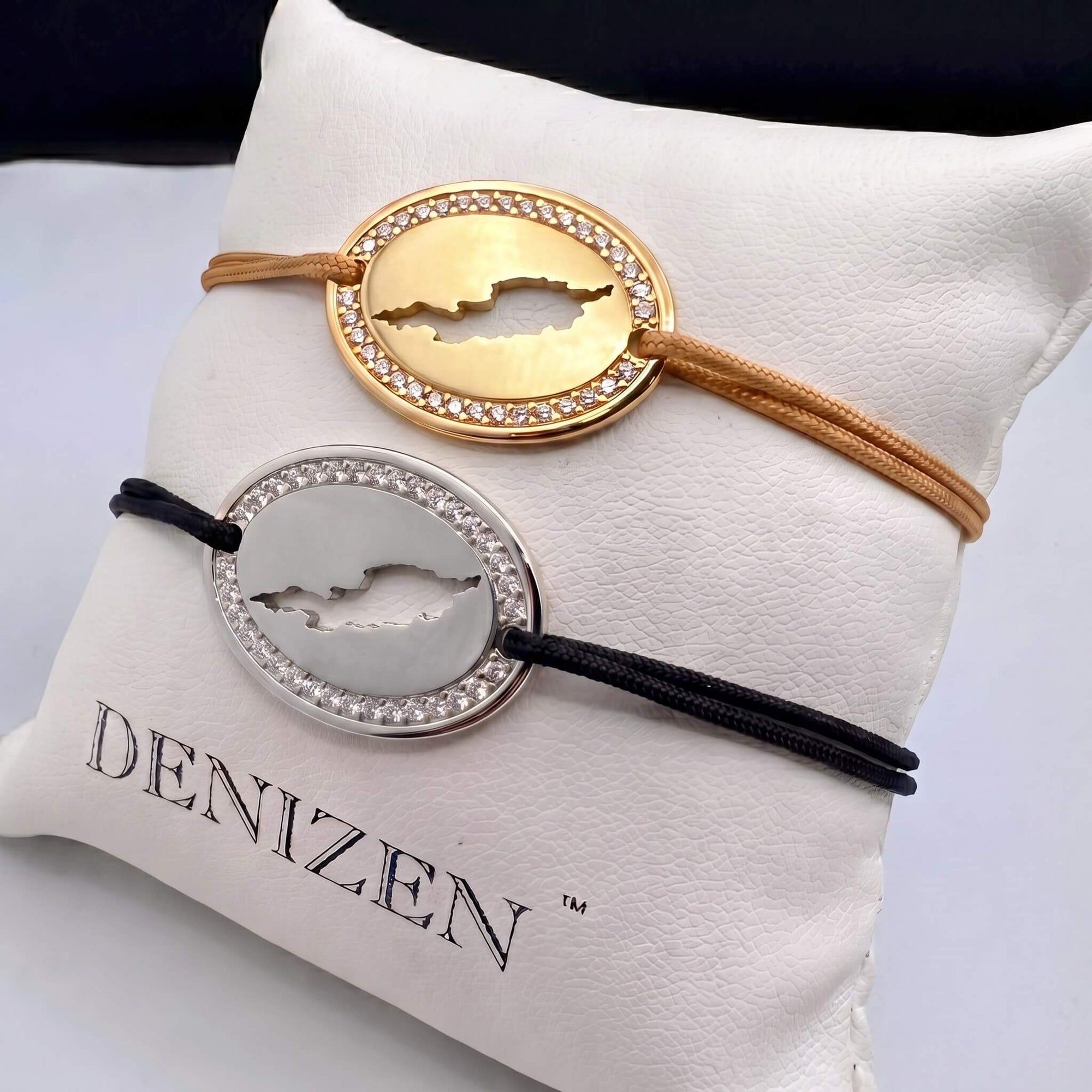 Two oval cord bracelets with a cut-out map of Anguilla and a crystal-studded border, displayed on a white pillow branded DENIZEN. The top bracelet is gold with a tan cord and the bottom bracelet is silver with a black cord