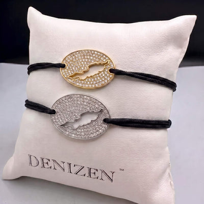 Two oval pendant bracelets with black cords and pavé-set pendants, featuring a cut-out map of Anguilla, one gold-toned and one silver-toned, displayed on a white cushion branded DENIZEN.