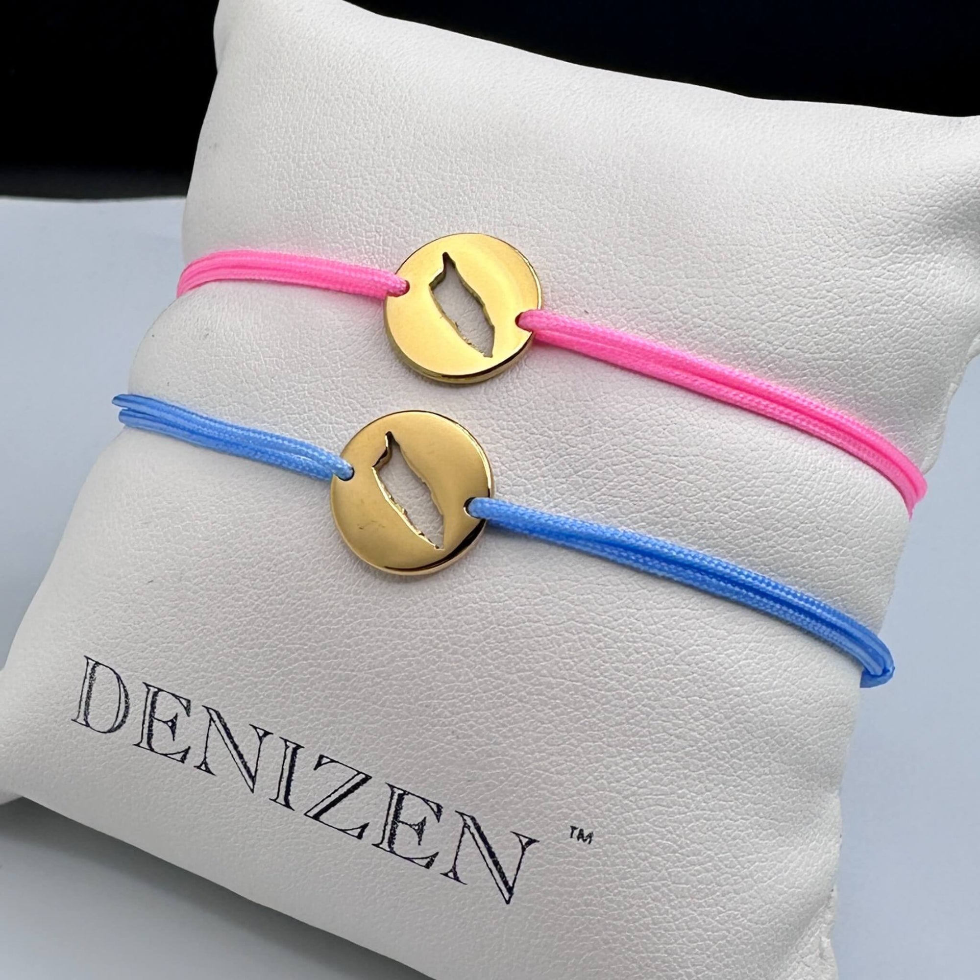 Two DENIZEN gold bracelets with an Aruba map cutout on pink and blue strings