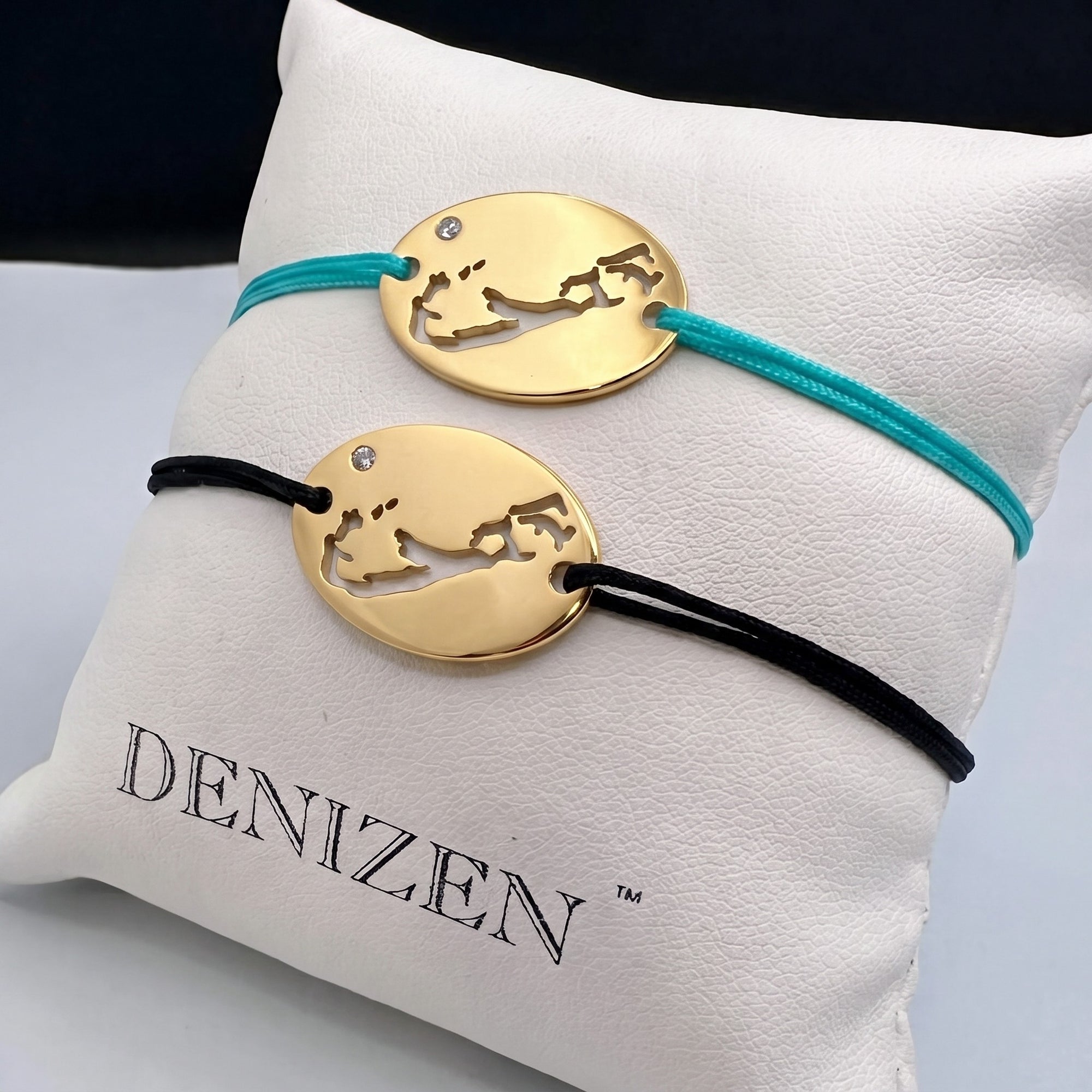 Two oval gold bracelet with cut-out maps of Bermuda on turquoise and black strings featuring a small clear stone displayed on a DENIZEN-branded white pillow.