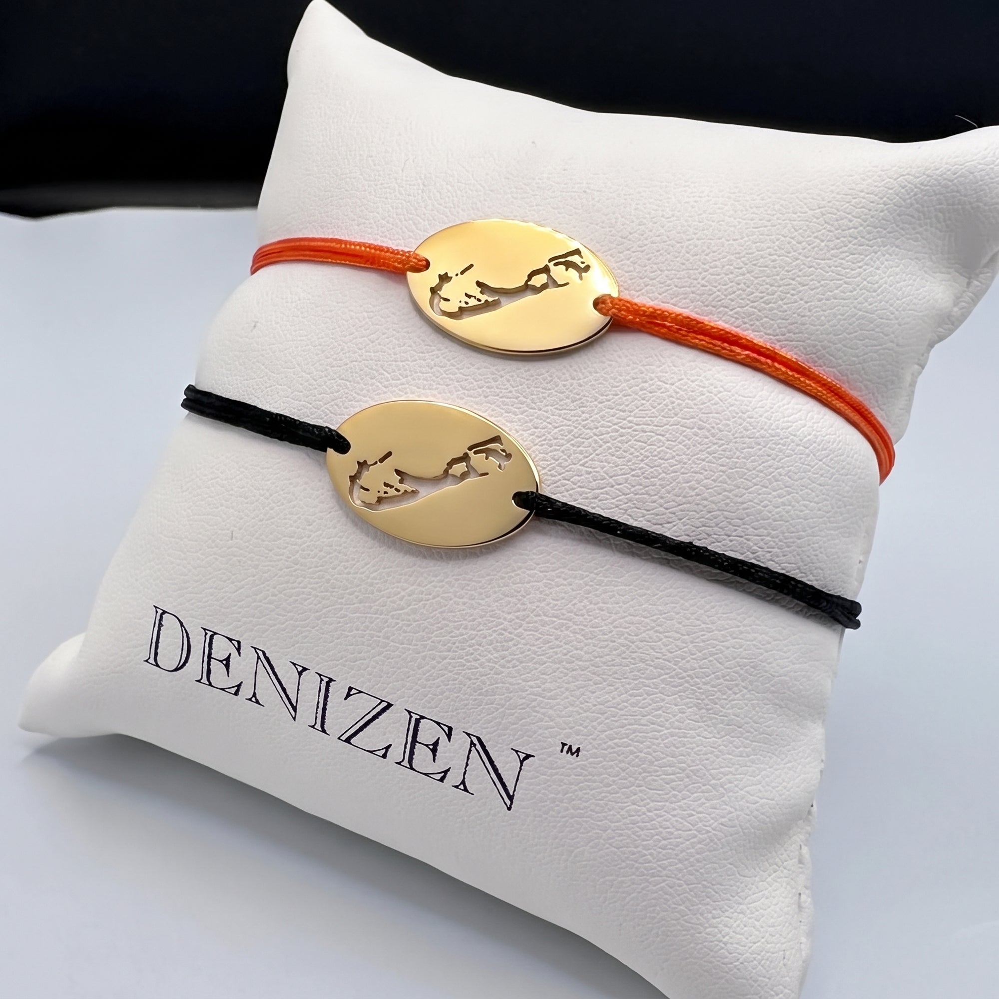 Two oval gold bracelet with cut-out maps of Bermuda on orange and black strings displayed on a DENIZEN-branded white pillow.