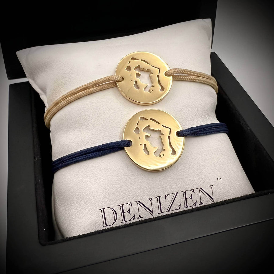 Two gold Denizen bracelets with a map cutout of Bora Bora displayed on a white cushion inside a black box. The top bracelet has a beige cord and the bottom one a navy cord.
