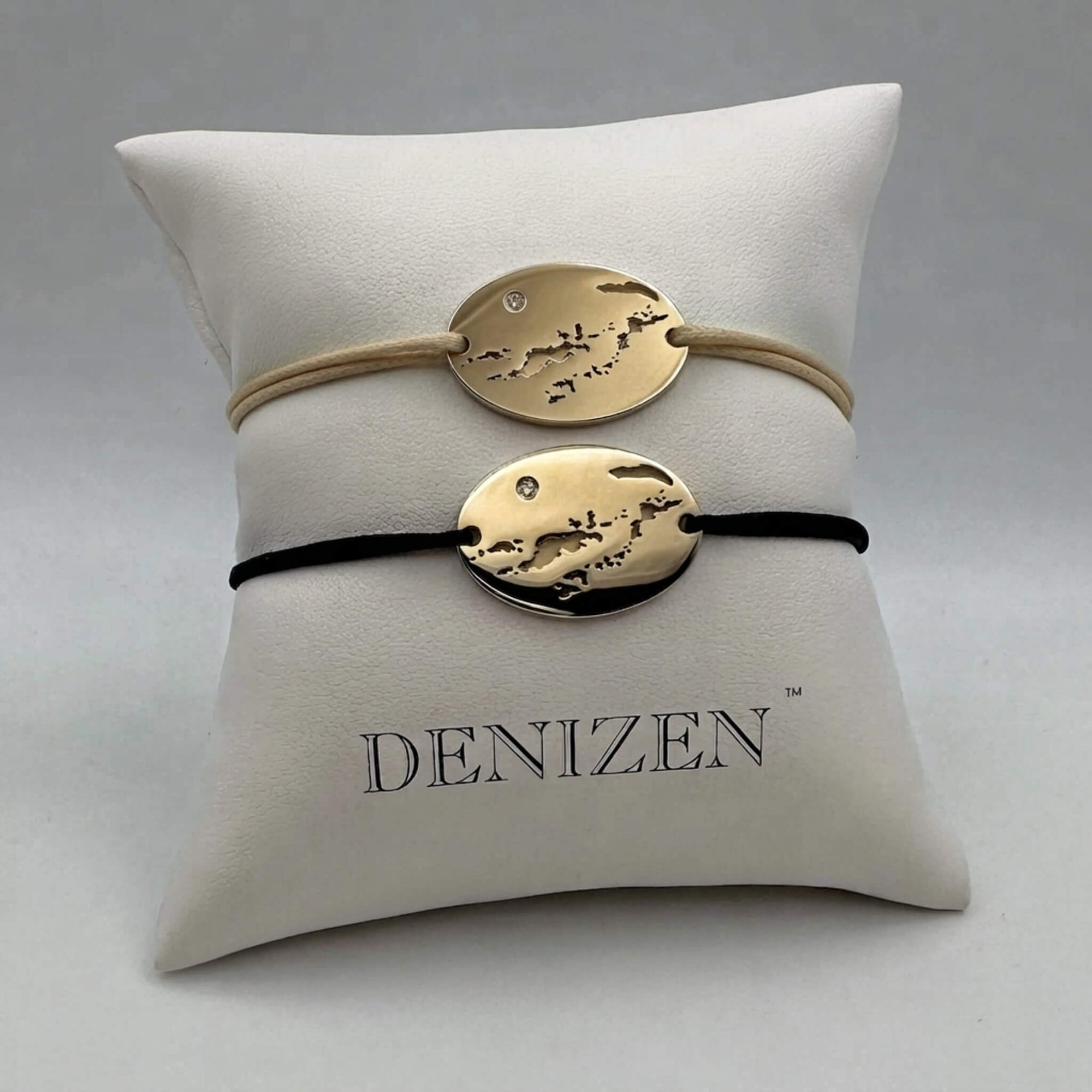 Two oval gold bracelets with a cut-out map of the BVI, one on a beige cord and one on a black cord, displayed on a white cushion branded DENIZEN.