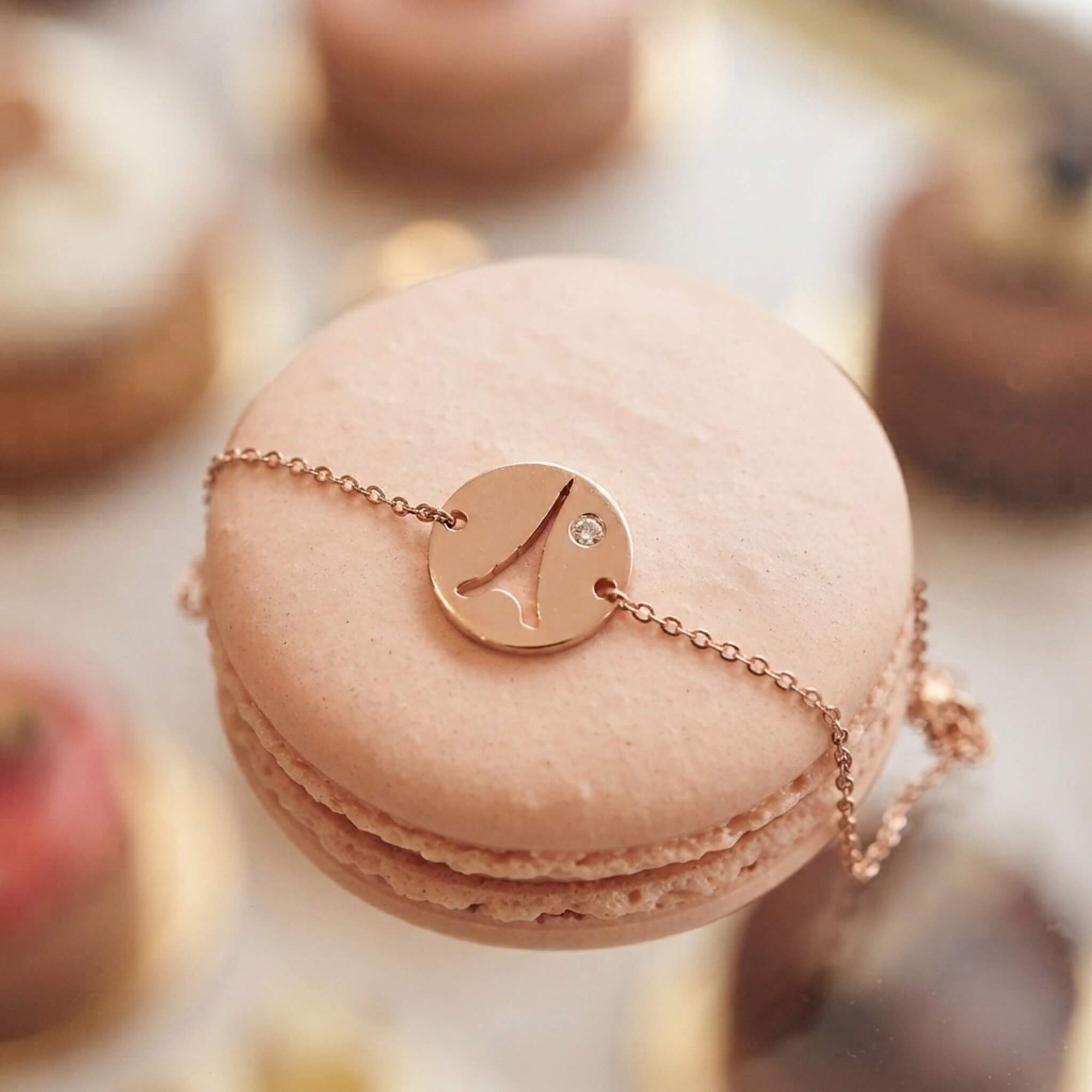 A close-up of a pale pink macaron decorated with a delicate rose gold chain bracelet featuring a cutout of the Eiffel Tower with a small clear stone, set against a soft-focus background of other macarons.