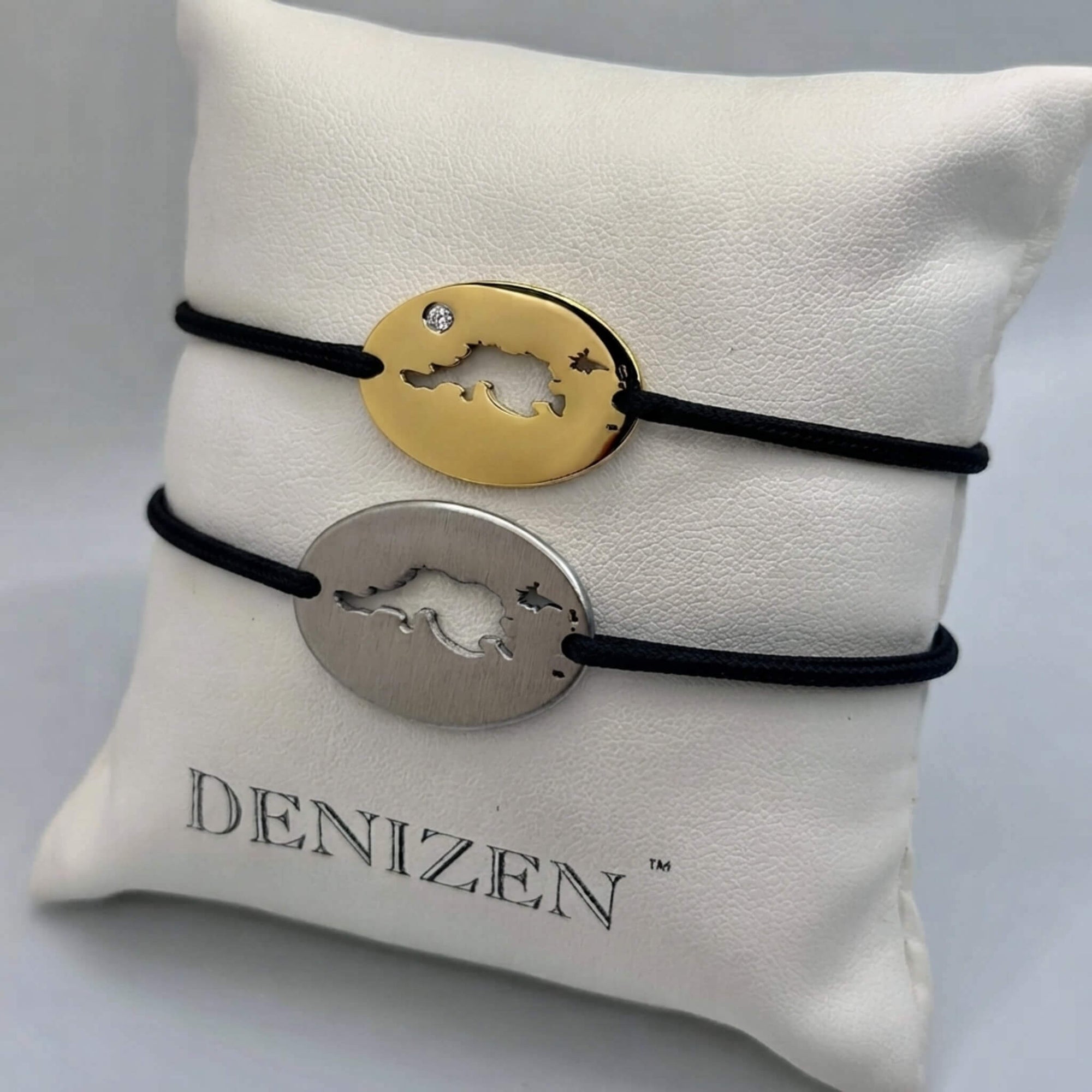 Two oval charm bracelets with black cords displayed on a white branded jewelry pillow. One bracelet has a gold-toned oval charm and the other has a silver-toned oval charm, both featuring a cutout design of Jost Van Dyke map and a small clear stone on the gold piece.