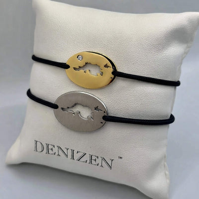 Two oval charm bracelets with black cords displayed on a white branded jewelry pillow. One bracelet has a gold-toned oval charm and the other has a silver-toned oval charm, both featuring a cutout design of Jost Van Dyke map and a small clear stone on the gold piece.