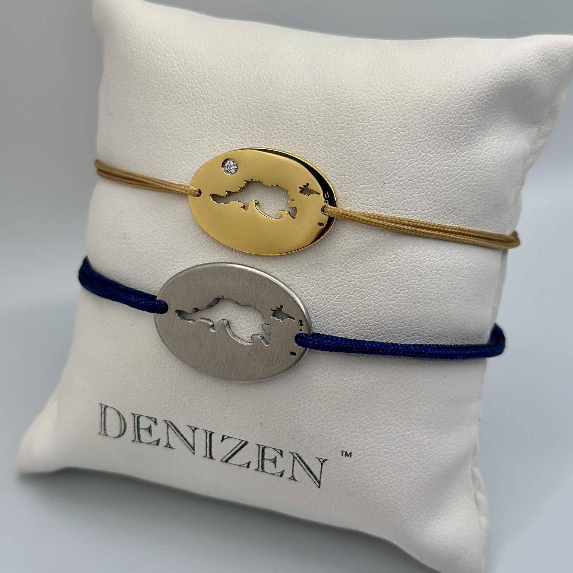 Two oval pendant bracelets displayed on a white DENIZEN branded pillow. The top bracelet is gold with a small clear stone on a tan cord. The bottom one is in brushed steel on a navy cord. Both feature a cut-out  map of Jost Van Dyke.