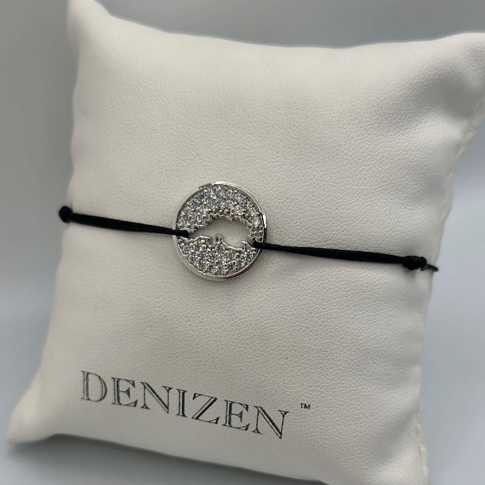 A black cord bracelet with a silver circular charm encrusted with clear stones and featuring a cut-out map of Jost Van Dyke, displayed on a white textured cushion printed with the brand name DENIZEN.