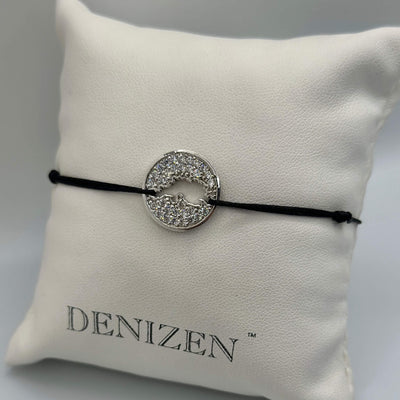 A black cord bracelet with a silver circular charm encrusted with clear stones and featuring a cut-out map of Jost Van Dyke, displayed on a white textured cushion printed with the brand name DENIZEN.