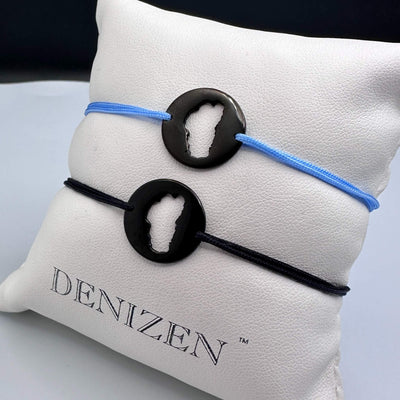 Two black circular pendant bracelets with cutouts of Lake Tahoe, one on a light blue cord and one on a black cord, displayed on a white branded pillow that reads DENIZEN