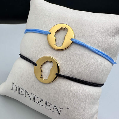 Two minimalist DENIZEN cord bracelets with polished gold disc pendants with cutouts of Lake Tahoe kare displayed on a white textured cushion branded DENIZEN. The top bracelet has a light blue cord, and the bottom has a black cord.
