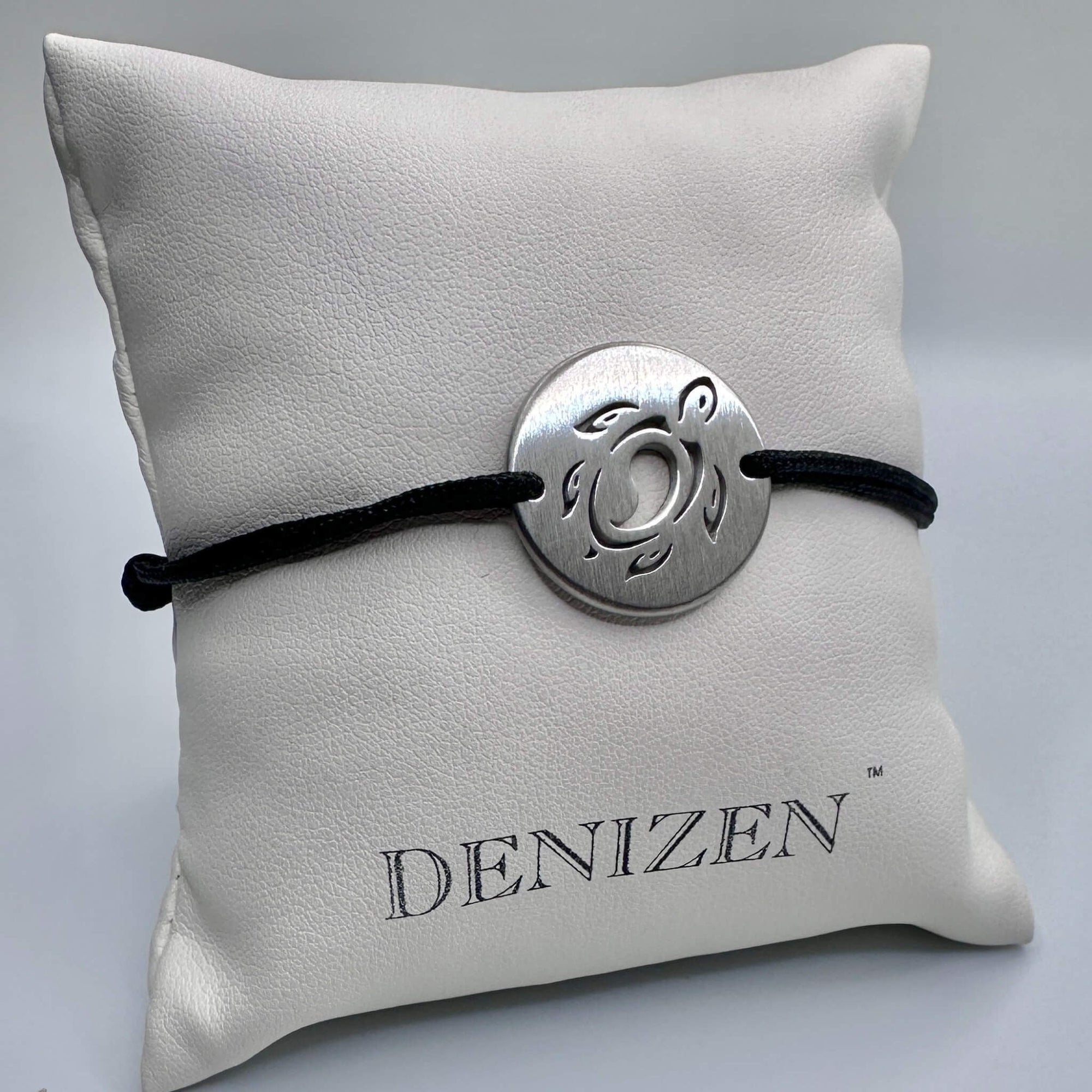 A silver circular pendant with a sea turtle cutout on a black cord bracelet, displayed on a white textured pillow with the brand name 'DENIZEN' printed on it.