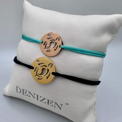 Two minimalist cord bracelets with circular metal charms displayed on a white cushion. The top bracelet has a turquoise cord and a rose gold charm, while the bottom bracelet has a black cord and a gold charm. Both have a sea turtle cutout design and a small clear  stone.
