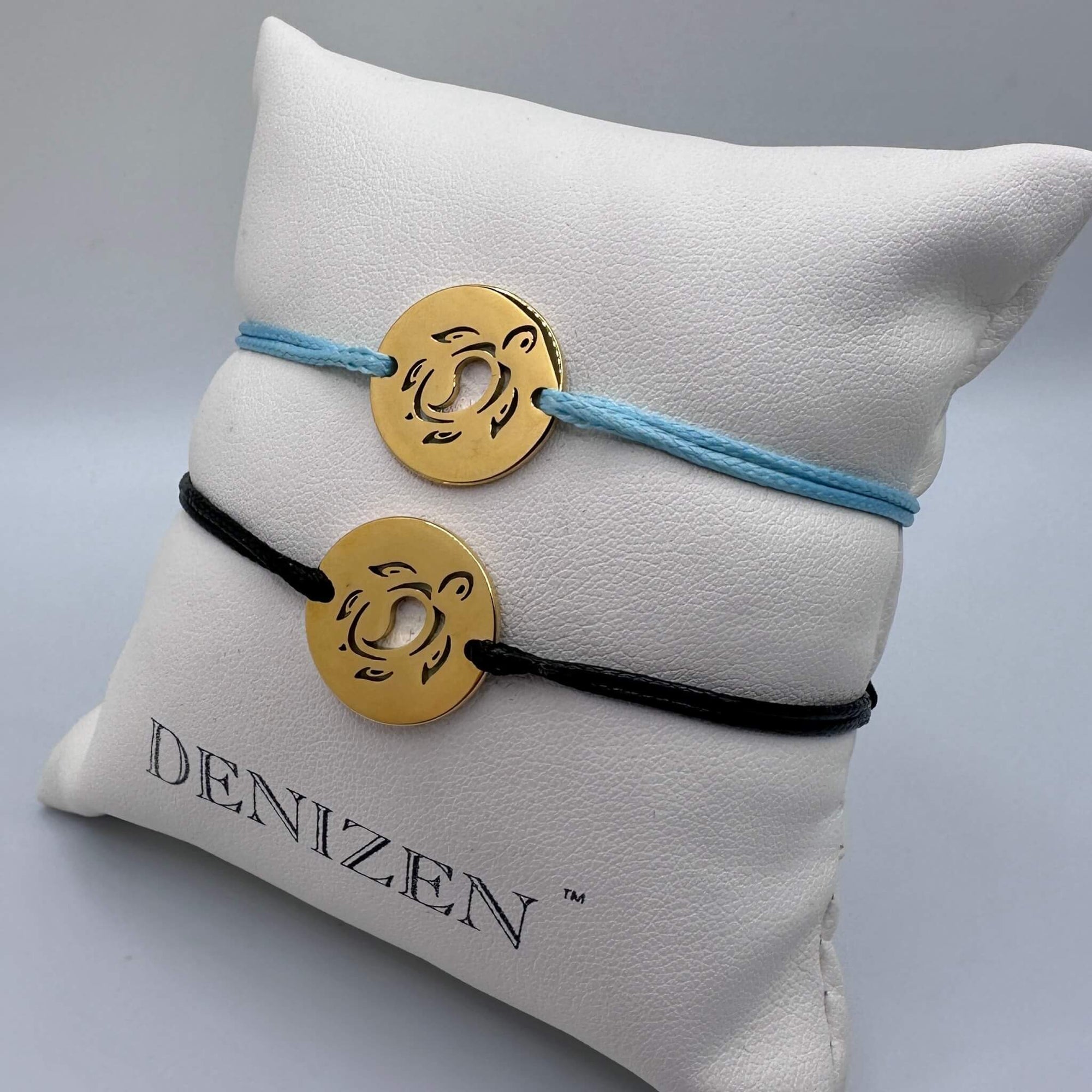 Two Denizen brand cord bracelets displayed on a white branded pillow. One bracelet is light blue with a gold circular medallion, and the other is black with a matching gold medallion, both with a sea turtle cutout design.