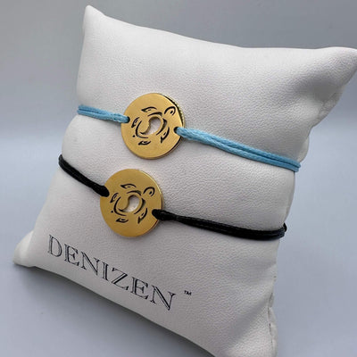 Two Denizen brand cord bracelets displayed on a white branded pillow. One bracelet is light blue with a gold circular medallion, and the other is black with a matching gold medallion, both with a sea turtle cutout design.