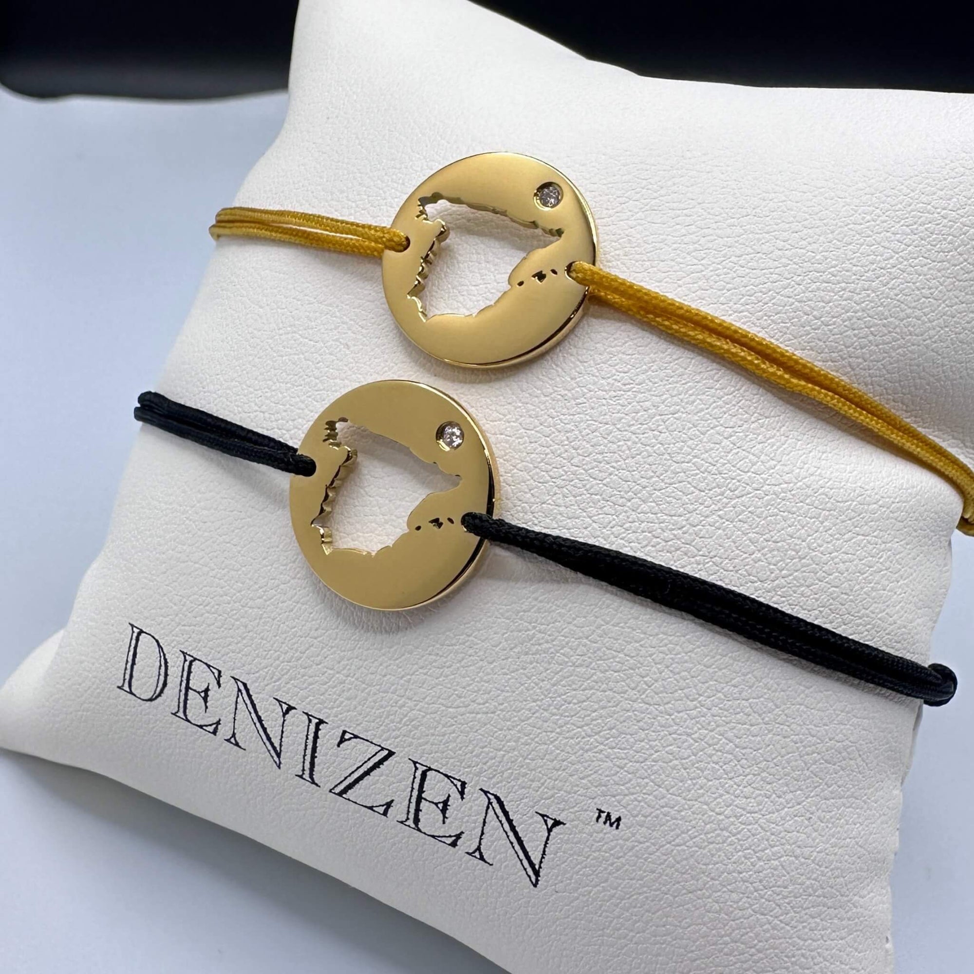 Two Denizen gold bracelets with a cut-out map if Spain displayed on a white branded cushion. One bracelet is on a beige string, the other on a black string.