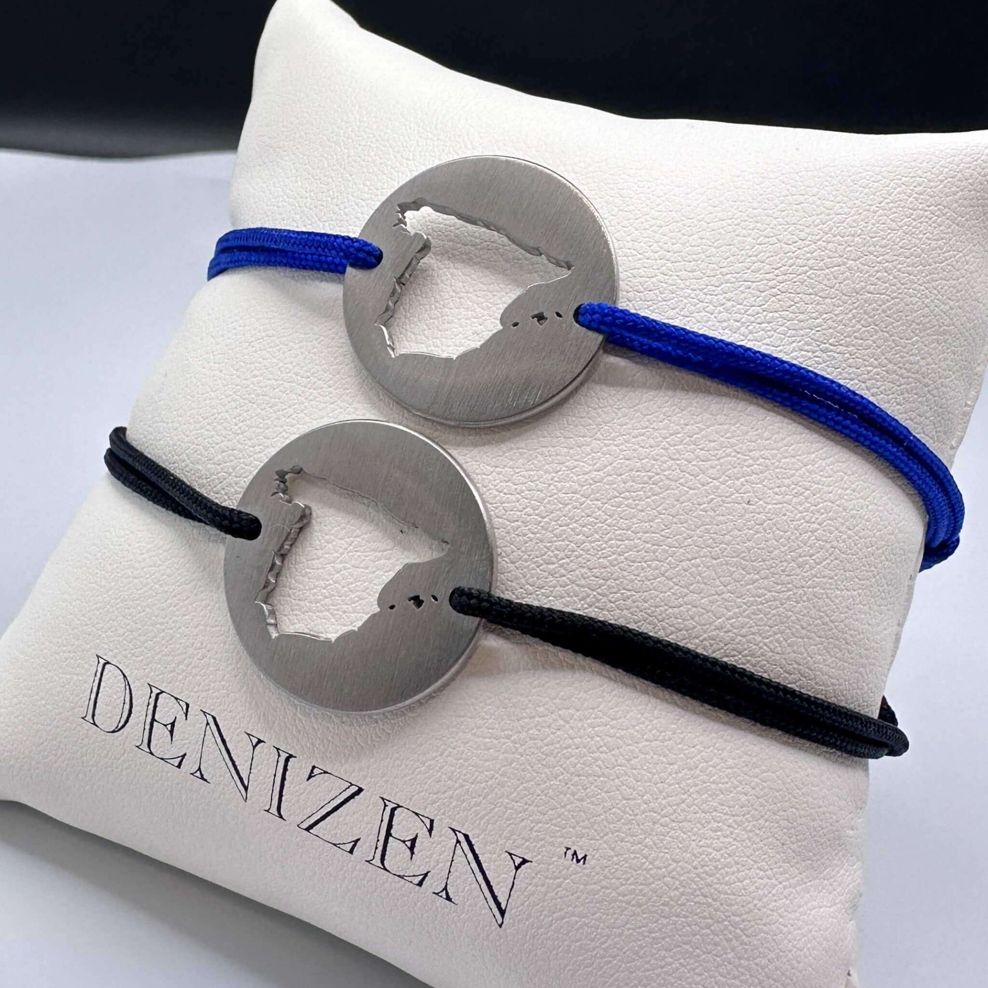 Two DENIZEN bracelets in brushed steel with the cut-out map of Spain, displayed on a white branded cushion. One bracelet has a blue cord and the other has a black cord.