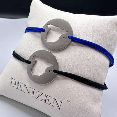 Two DENIZEN bracelets in brushed steel with the cut-out map of Spain, displayed on a white branded cushion. One bracelet has a blue cord and the other has a black cord.