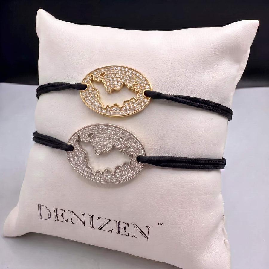 A round gold pendant necklace covered in clear pavé stones, featuring a cutout of St-Barth map, resting on sand.