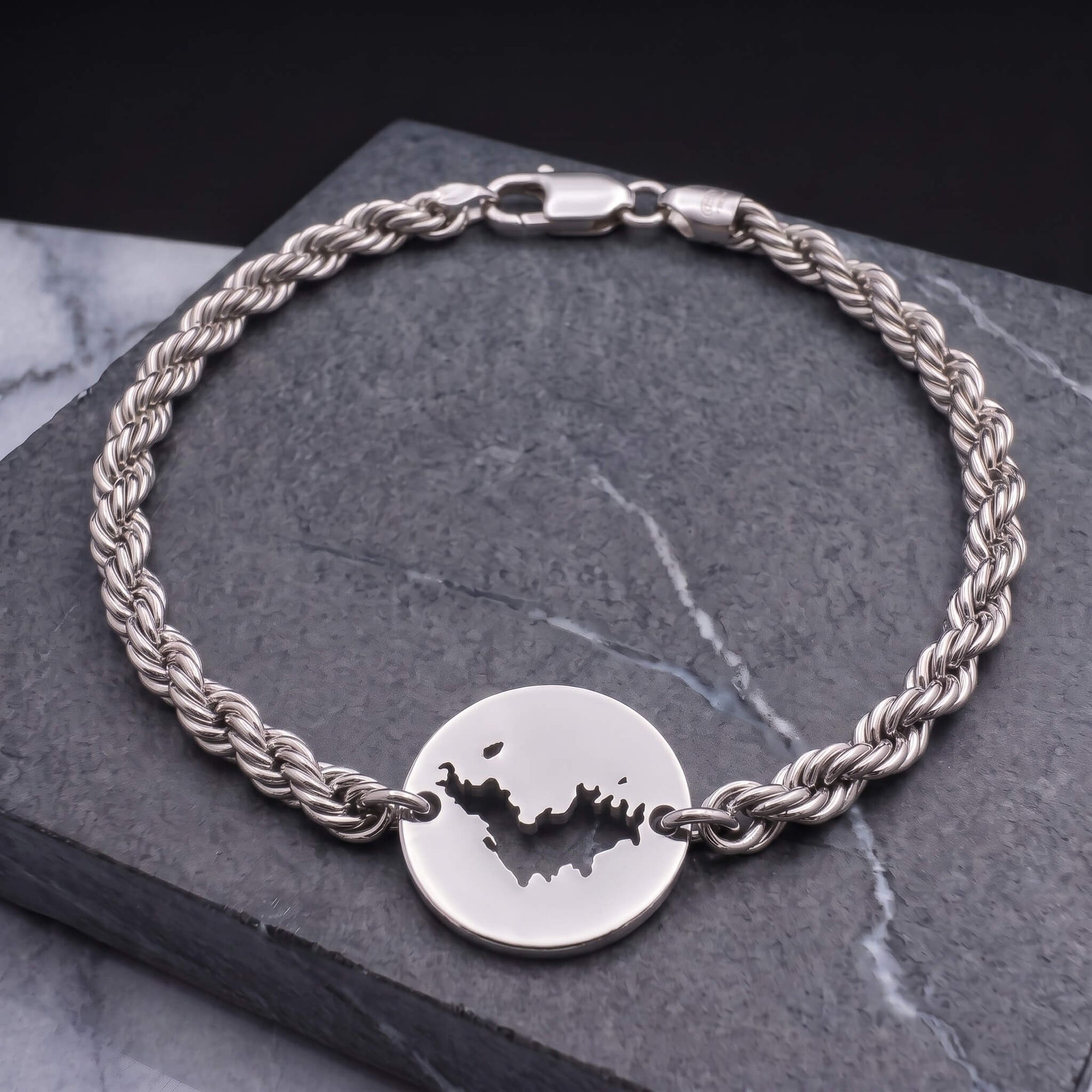 A silver-toned twisted rope chain bracelet with a round disc charm featuring a cutout map of St-Barth, displayed on a dark gray stone slab.