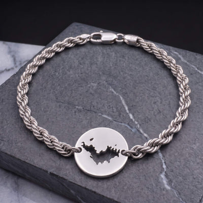A silver-toned twisted rope chain bracelet with a round disc charm featuring a cutout map of St-Barth, displayed on a dark gray stone slab.