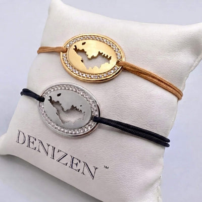 Two oval bracelets with a cutout map of St-Barth and an outer ring of clear stones, displayed on a white cushion branded DENIZEN. The top gold bracelet has a tan cord. and the bottom silver bracelet has a black cord.