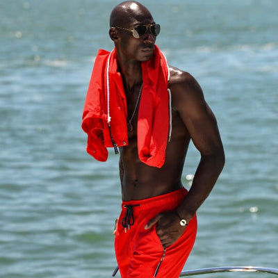 A shirtless man on board a boat wearing bright red drawstring shorts and a matching red zip-up jacket draped over his shoulders, and a silver DENIZEN bracelet with a cutout map of St Barth.