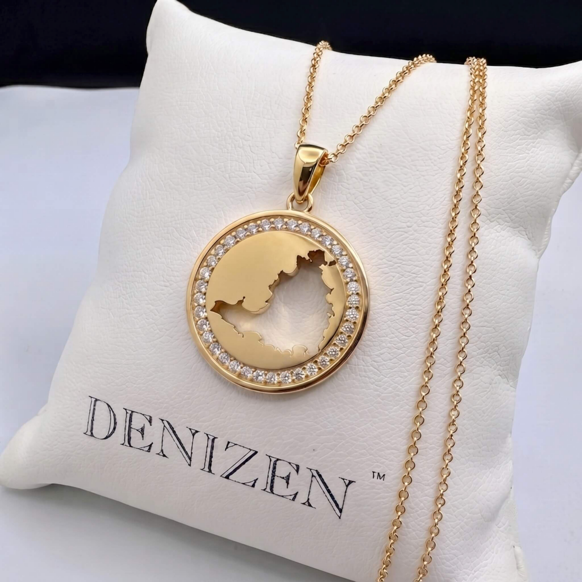A gold pendant necklace with a cutout of St. Maarten map surrounded by a halo of clear stones, displayed on a white cushion printed with the brand name 'DENIZEN'.