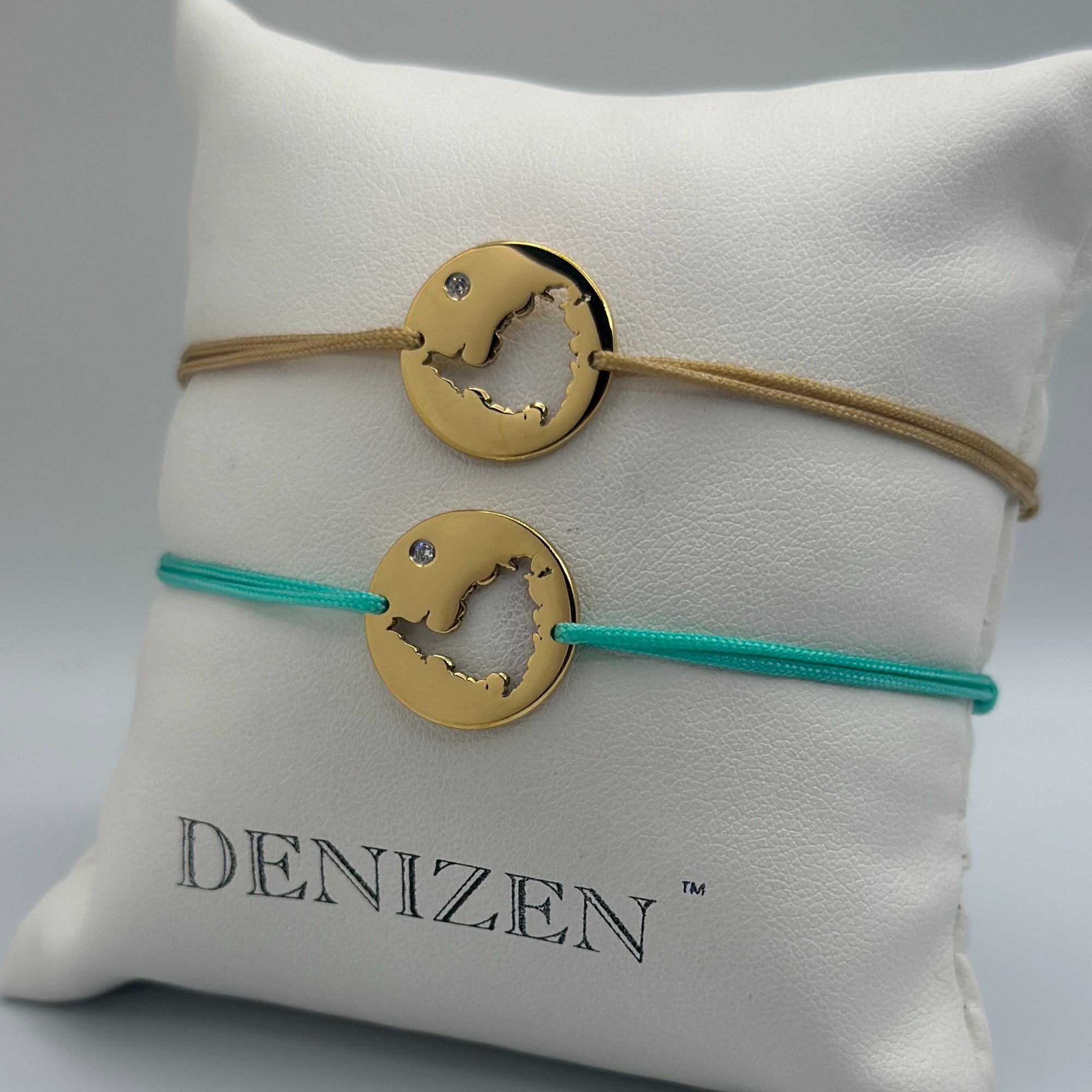 Two gold-toned circular pendant bracelets with a cut-out map of St. Maarten and small clear stones, displayed on a white cushion. One bracelet has a tan cord and the other has a turquoise cord.