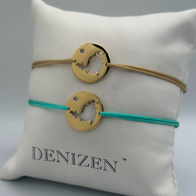 Two gold-toned circular pendant bracelets with a cut-out map of St. Maarten and small clear stones, displayed on a white cushion. One bracelet has a tan cord and the other has a turquoise cord.