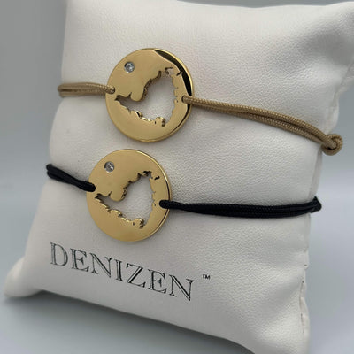 Two gold-toned cord bracelets with cutouts of St. Maarten map and small clear stones, displayed on a white branded Denizen cushion. One bracelet has a tan cord and the other has a black cord.