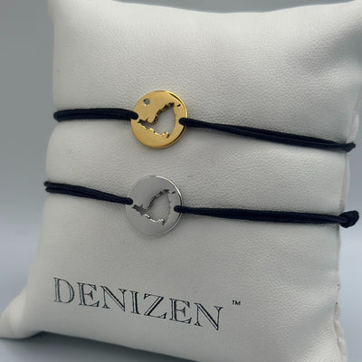 Two Denizen brand cord bracelets displayed on a white branded pillow. One bracelet is gold with a small clear stone and the other is silver-tone . Both bracelets a re on black cord and feature a cut-out map of St. Maarten.