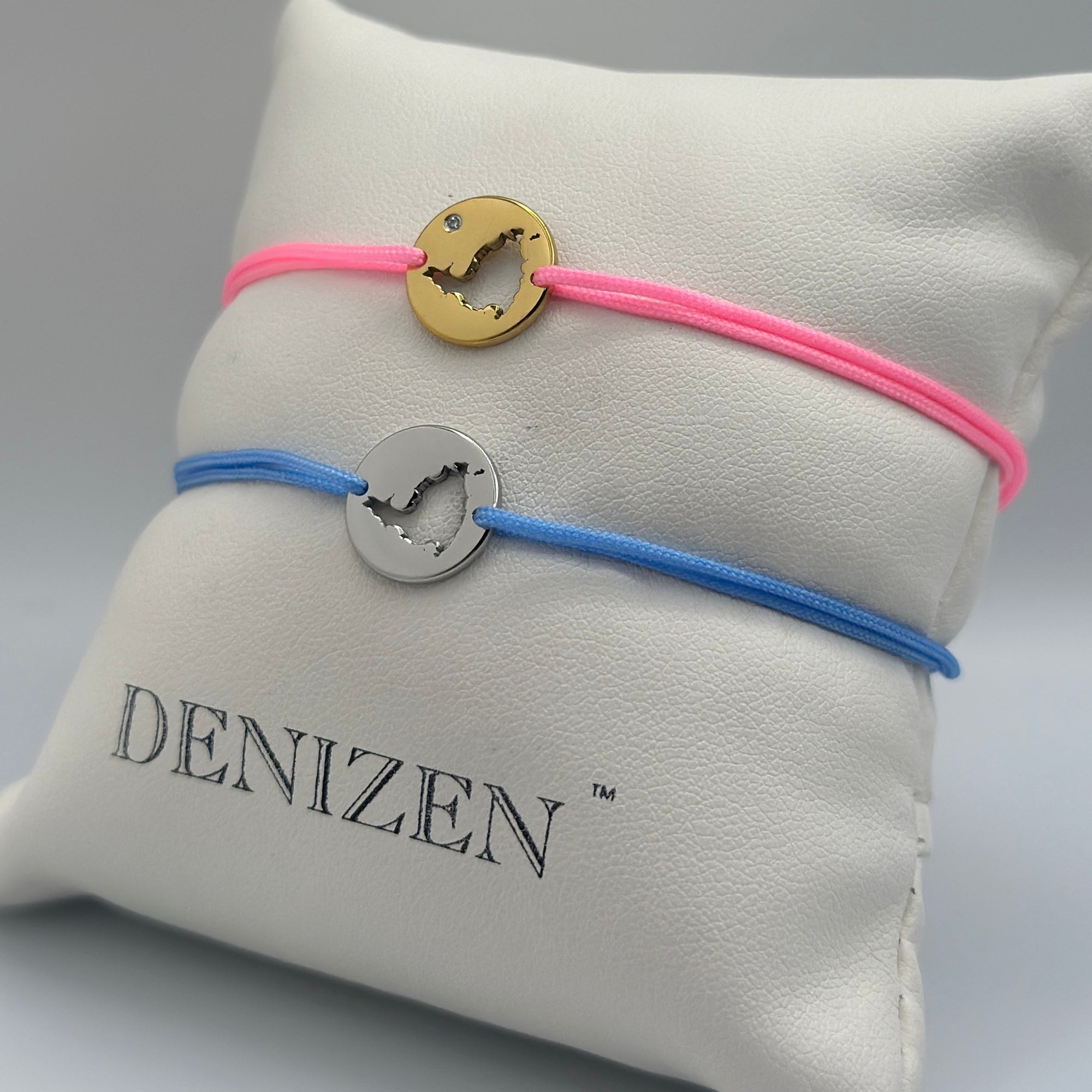 Two Denizen brand cord bracelets displayed on a white branded pillow. One bracelet is pink with a gold-tone circular disc charm and a clear stone, and the other is blue with a silver-tone circular disc charm. Both bracelets feature a cut-out map of St. Maarten.