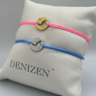 Two Denizen brand cord bracelets displayed on a white branded pillow. One bracelet is pink with a gold-tone circular disc charm and a clear stone, and the other is blue with a silver-tone circular disc charm. Both bracelets feature a cut-out map of St. Maarten.