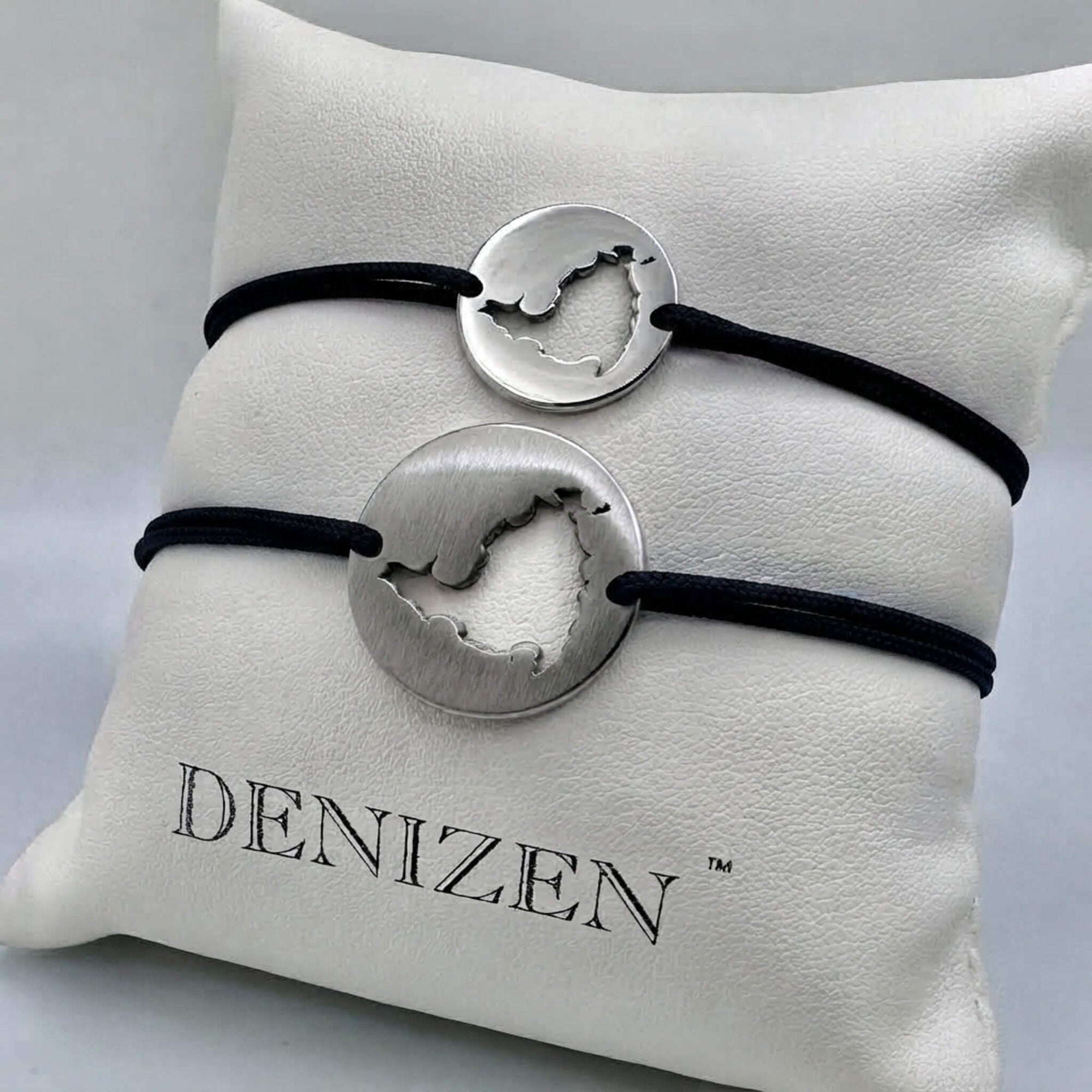 Two silver-toned cord bracelets with cutouts of St. Maarten map, displayed on a white branded Denizen cushion. One polished bracelet has a red cord and the other is brushed on navy cord.