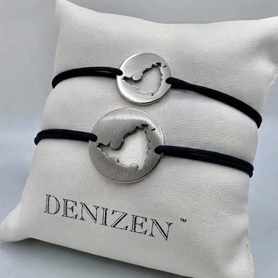 Two silver-toned cord bracelets with cutouts of St. Maarten map, displayed on a white branded Denizen cushion. One polished bracelet has a red cord and the other is brushed on navy cord.