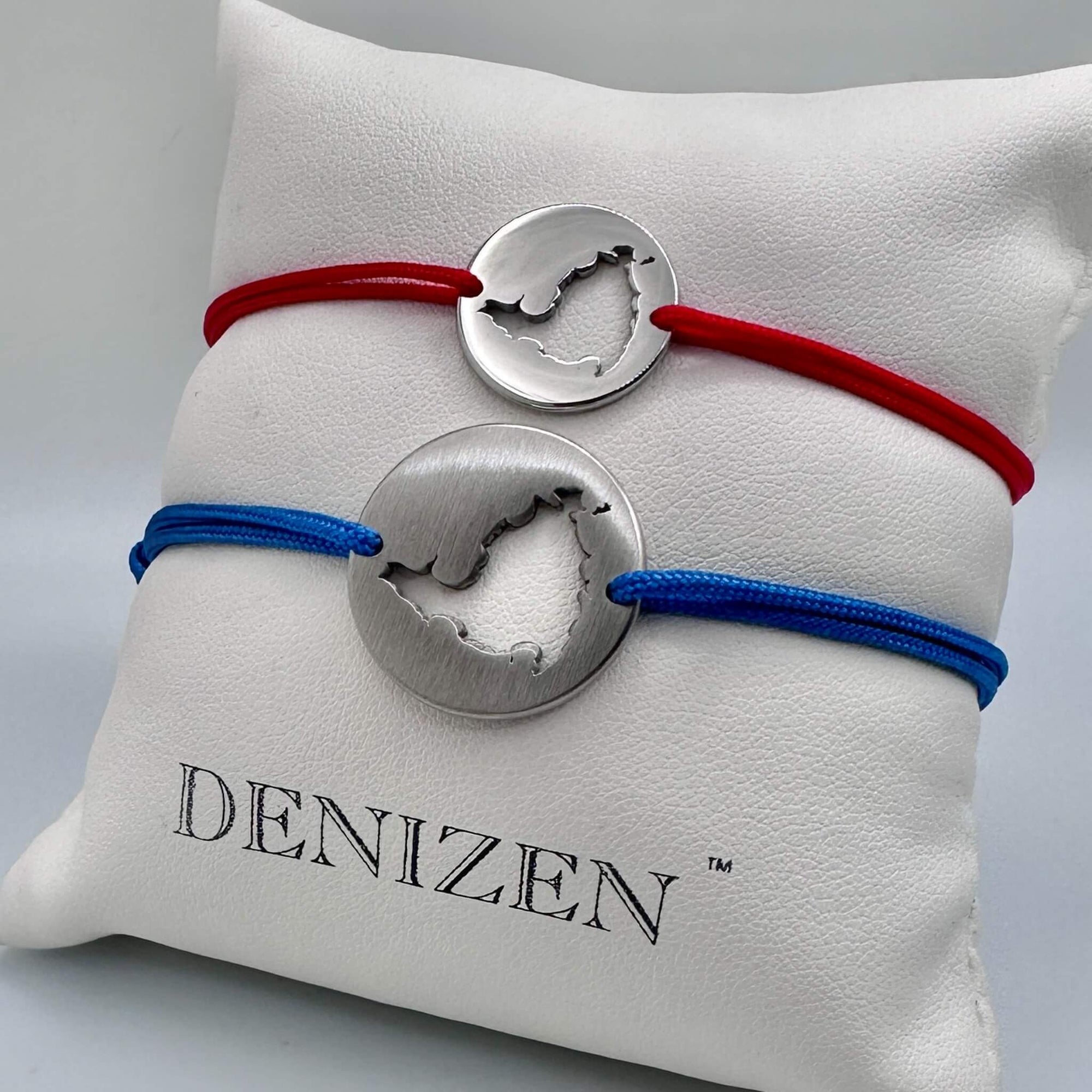 Two silver-toned cord bracelets with cutouts of St. Maarten map, displayed on a white branded Denizen cushion. One polished bracelet has a red cord and the other is brushed on navy cord.