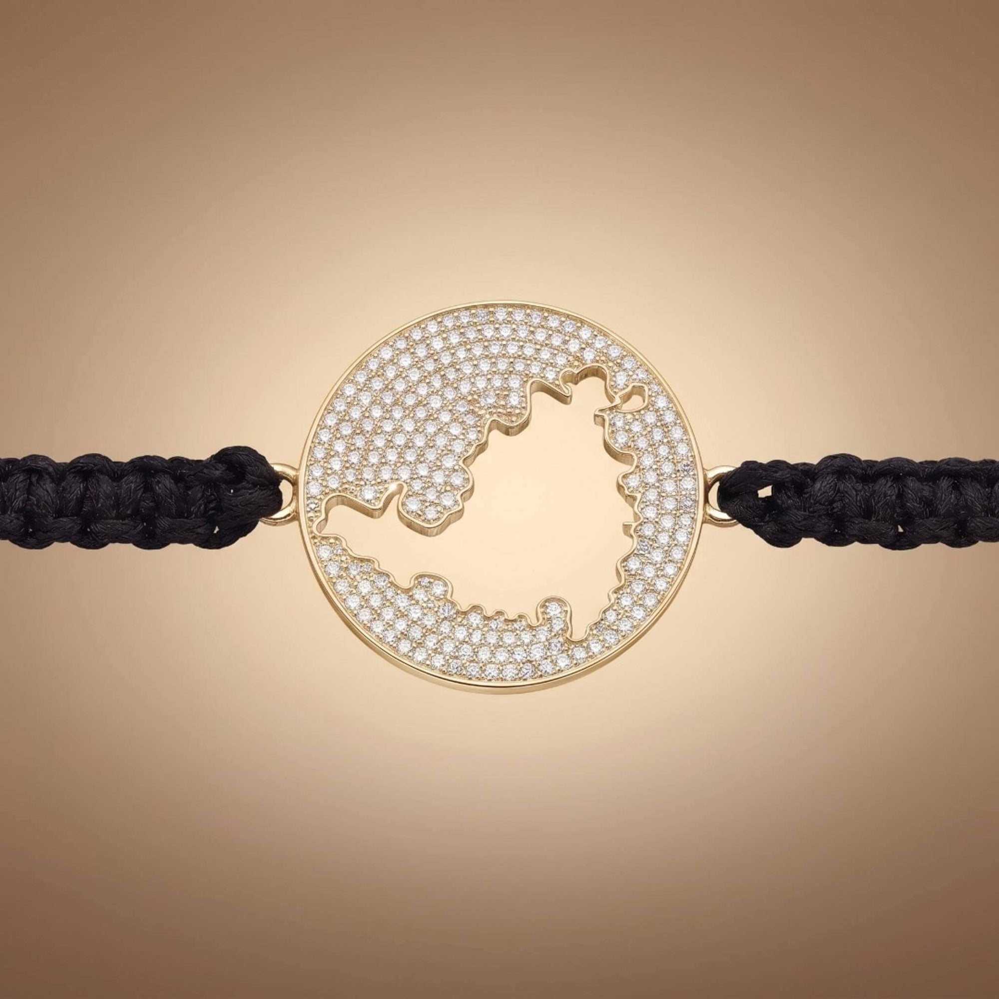 A gold round bracelet featuring a cutout map  of St Maarten embellished with small clear pavé stones, connected to a black braided cord on a warm beige gradient background.