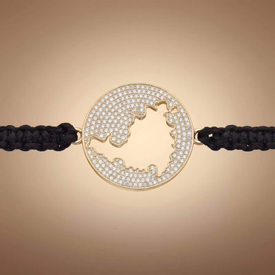 A gold round bracelet featuring a cutout map  of St Maarten embellished with small clear pavé stones, connected to a black braided cord on a warm beige gradient background.