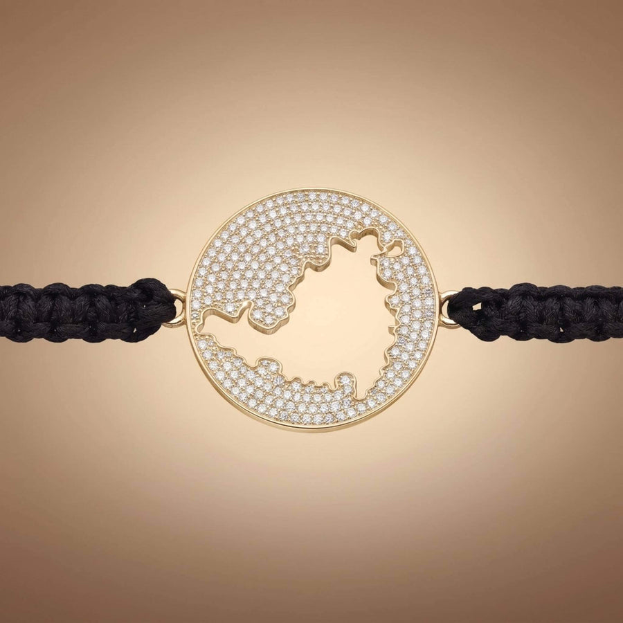 A gold round bracelet featuring a cutout map  of St Maarten embellished with small clear pavé stones, connected to a black braided cord on a warm beige gradient background.
