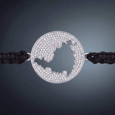 A silver round pendant bracelet with a cutout map of St Maarten and a diamond pave, set on a black braided cord bracelet against a dark gray gradient background.