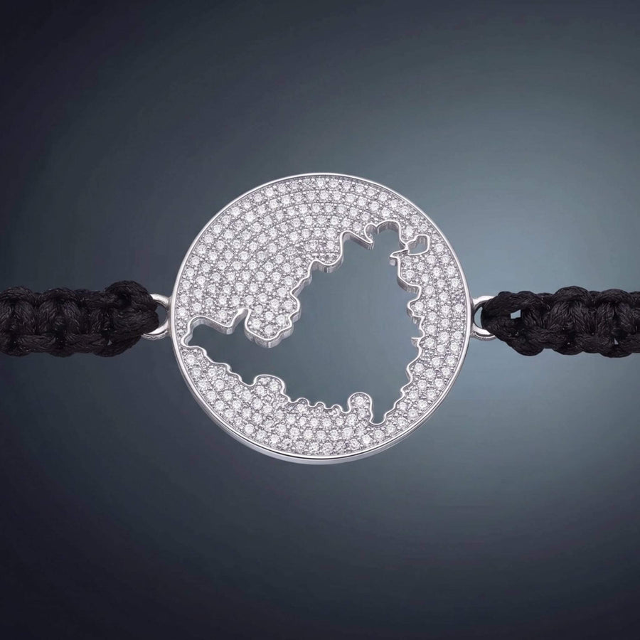 A gold round bracelet featuring a cutout map  of St Maarten embellished with small clear pavé stones, connected to a black braided cord on a warm beige gradient background.