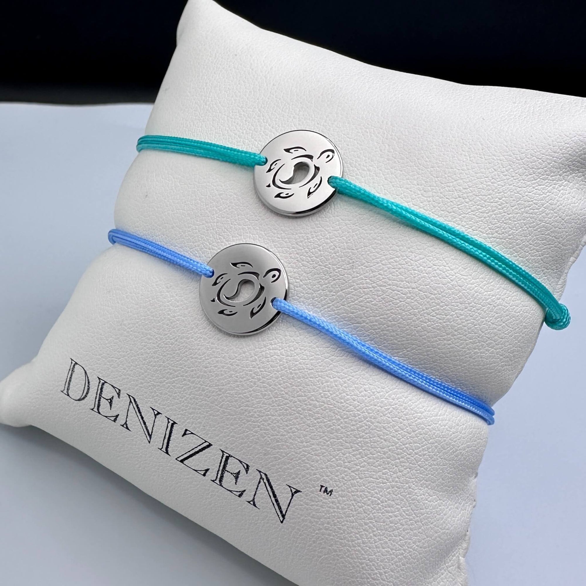 Two silver disc charms with a sea turtle cutout on thin cord bracelets, one in teal and one in light blue, displayed on a white textured cushion printed with the brand name 'DENIZEN'.
