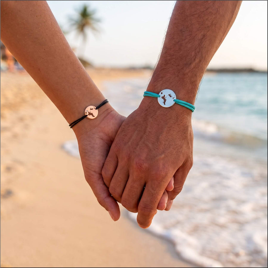 couple holding hands on a beach wearing DENIZEN bracelets on black and turquoise strings with a cutout map of Seychelles in rose gold and silver