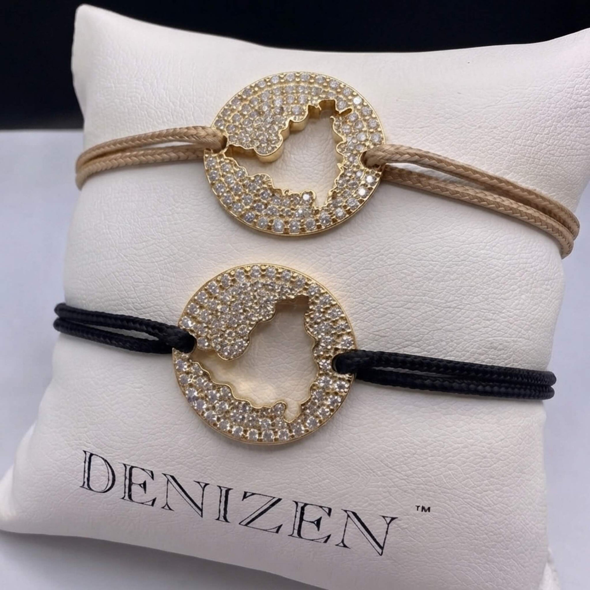 Two Denizen brand bracelets displayed on a white branded cushion. One bracelet has a beige cord and the other has a black cord, both featuring a gold-toned circular pendant encrusted with diamond pavé and cut out in the shape of the St. Maarten map.