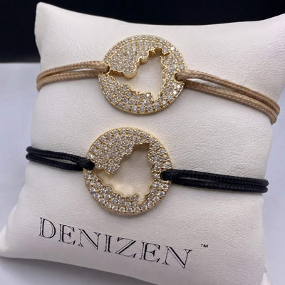 Two Denizen brand bracelets displayed on a white branded cushion. One bracelet has a beige cord and the other has a black cord, both featuring a gold-toned circular pendant encrusted with diamond pavé and cut out in the shape of the St. Maarten map.