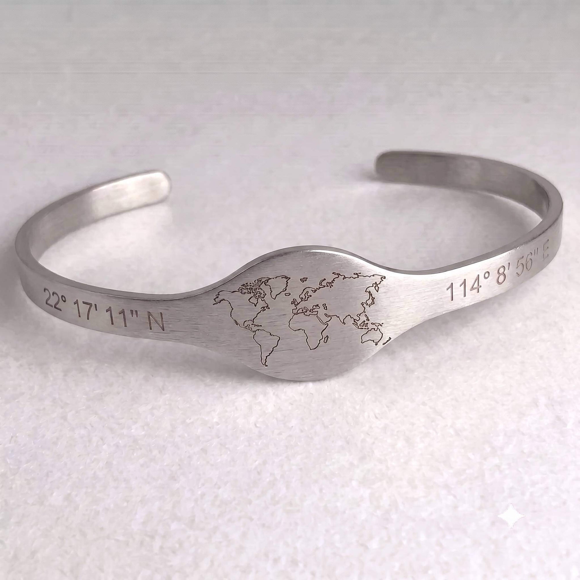 A brushed silver metal cuff bracelet featuring an engraved world map in the center and geographic coordinates on both ends, displayed on a light textured background.
